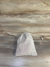 Top-down view of a beige drawstring pouch containing a 5-pack of pink baby mittens size 0-6 months on a wood surface