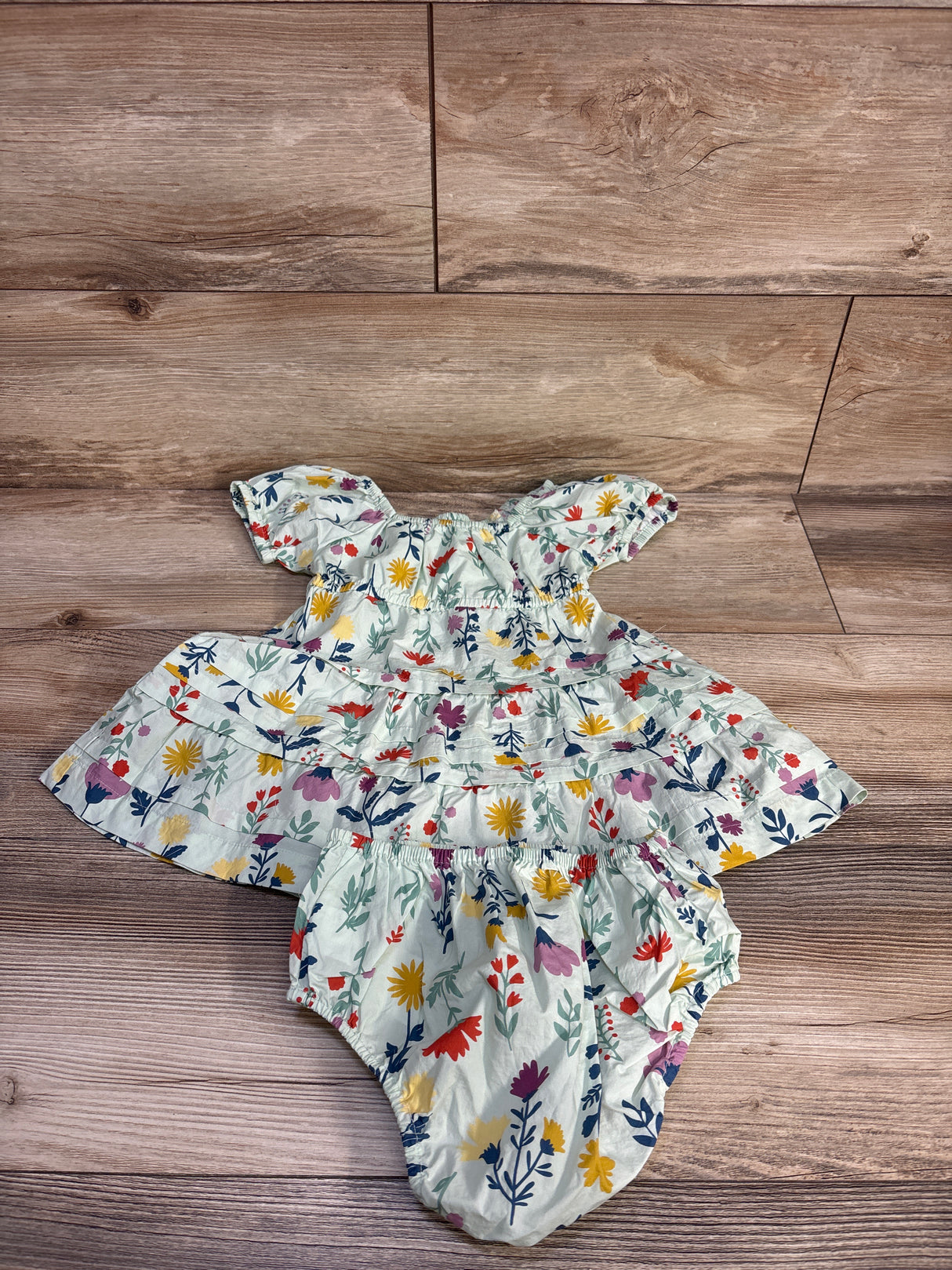 Flat lay of green floral cotton tiered dress with matching bloomers, puffed sleeves and tiered skirt on wood background
