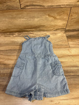 Flat-lay of blue chambray toddler romper showing ruffle shoulder straps, front patch pockets, and snap crotch on wood floor