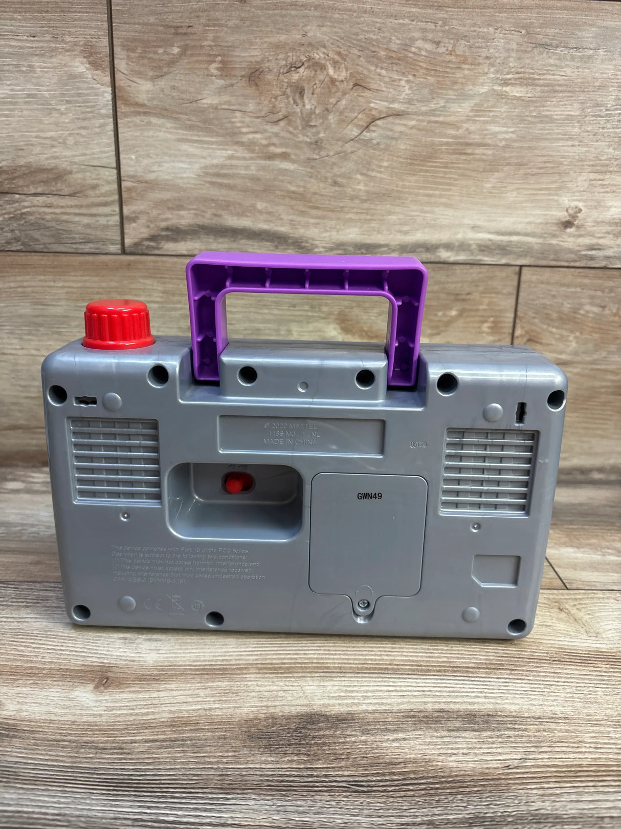 Back view of Fisher Price Laugh & Learn Busy Boombox with purple handle, red knob, and speaker grilles on wood-grain background