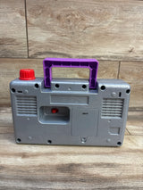 Back view of Fisher Price Laugh & Learn Busy Boombox with purple handle, red knob, and speaker grilles on wood-grain background