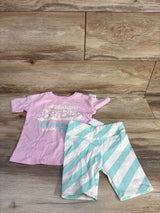 Flat lay of toddler two-piece: pink shirt with Malibu Barbie graphic and mint-and-white striped shorts on wood floor