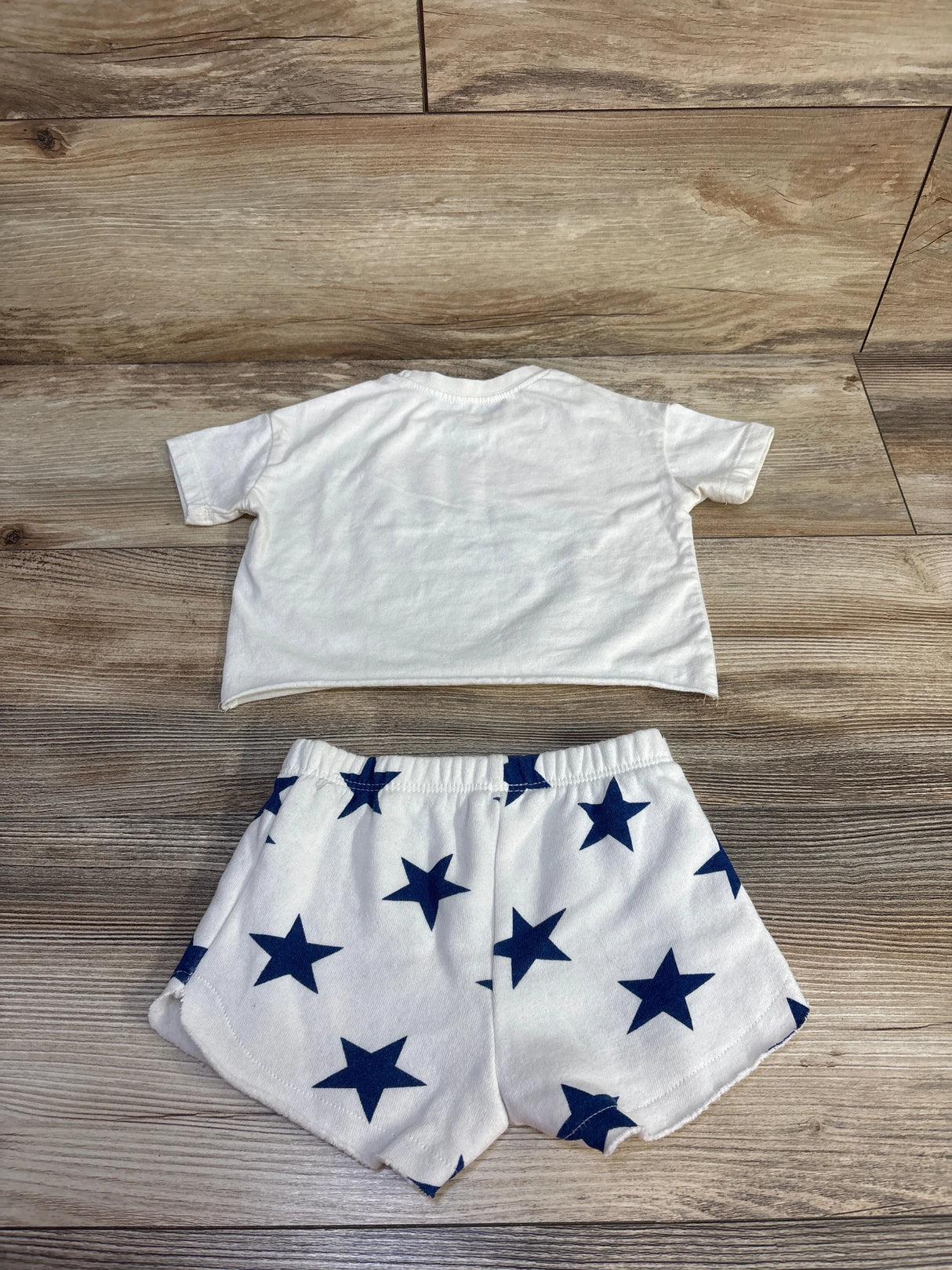 Flat lay of Grayson Mini 2-piece USA shirt and shorts showing back of ivory tee and white shorts with navy stars on wood