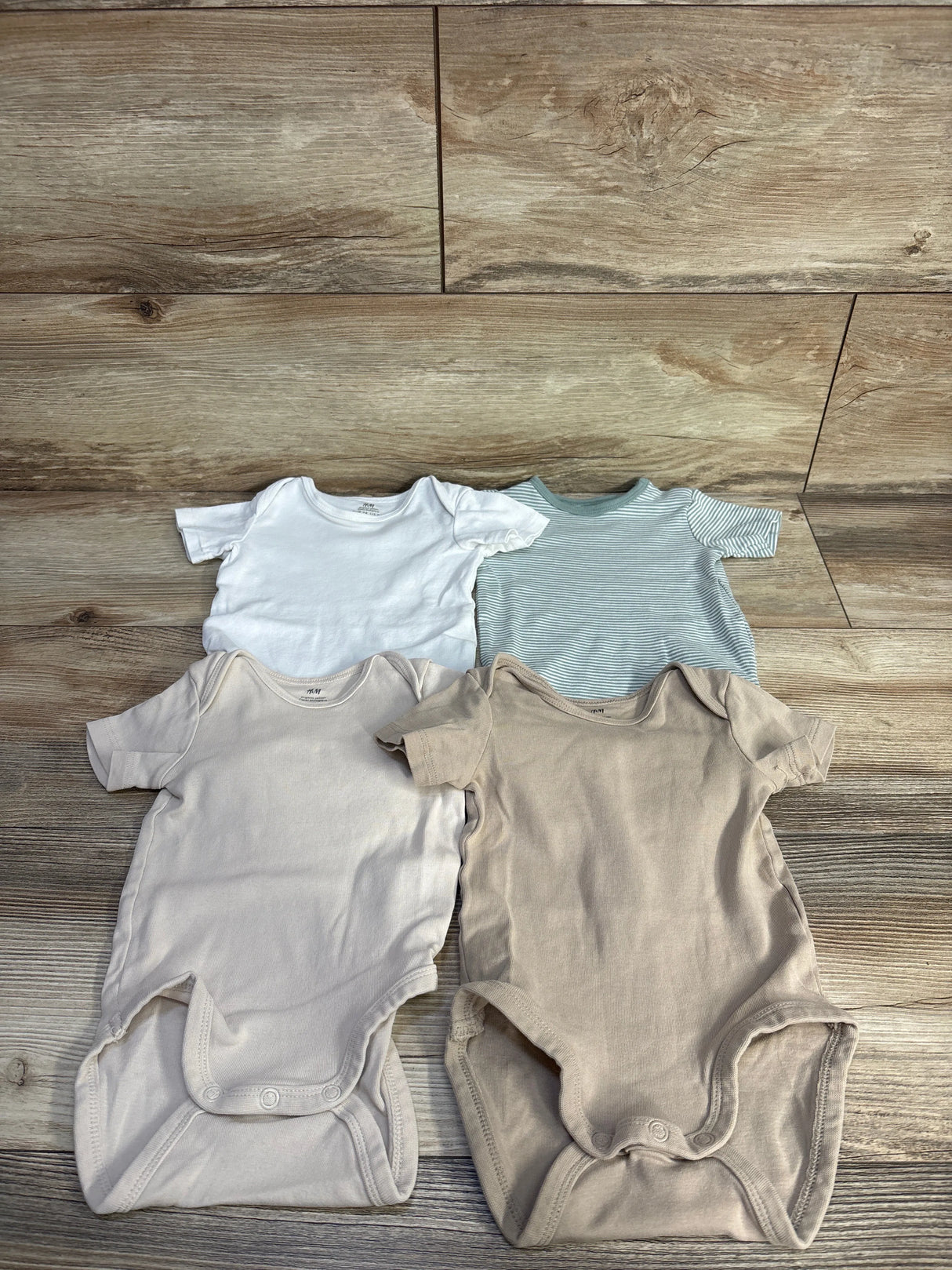 Flat lay of H&M organic 4-pack kimono bodysuits showcasing short sleeves, envelope necks, and snap crotches with wood floor background