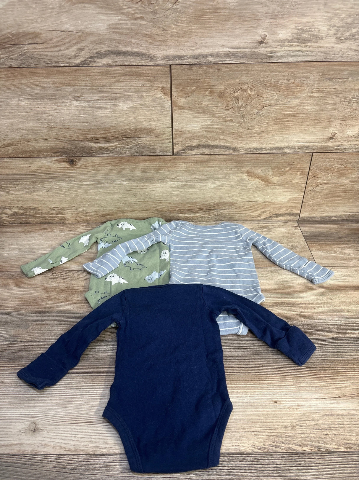 Flat lay of three long-sleeve baby bodysuits in navy, gray stripe, and green dinosaur print on wood background, size 3 months