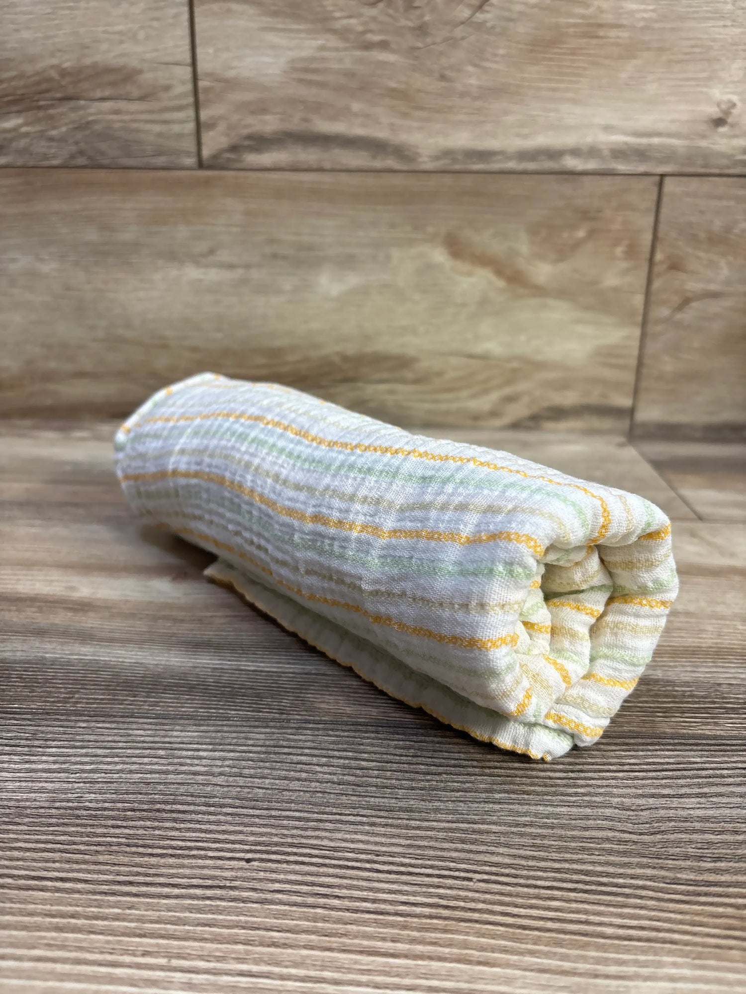 Angled view of Little Me striped muslin swaddle blanket showcasing white fabric with yellow, gray, and green stripes on wood
