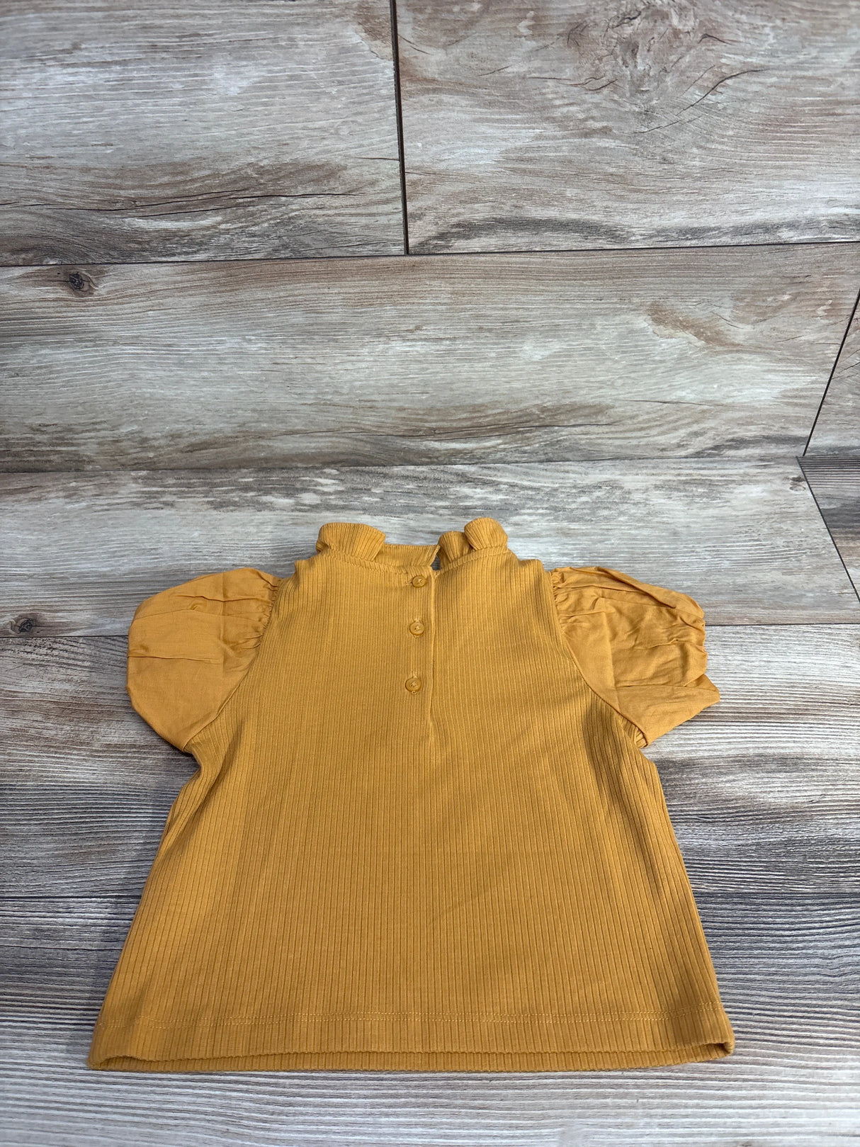 Mustard yellow toddler top with puff sleeves and three back buttons on wood background