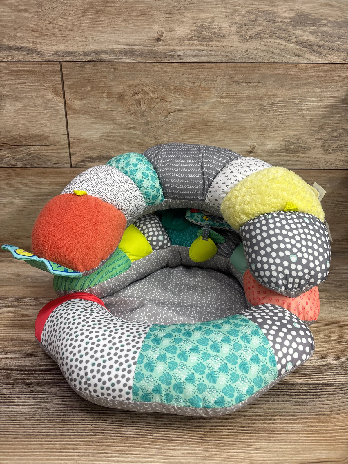 Top view of Infantino 2-in-1 Tummy Time and Seated Support showcasing colorful patterned cushions and attached toys on a wood floor