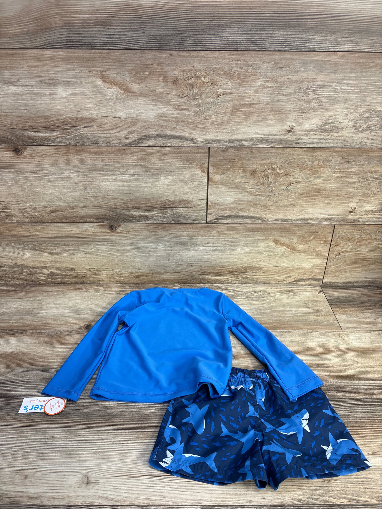 Flat lay of 2-piece shark-print rash guard set with blue long-sleeve top and shark-pattern swim shorts on wood background