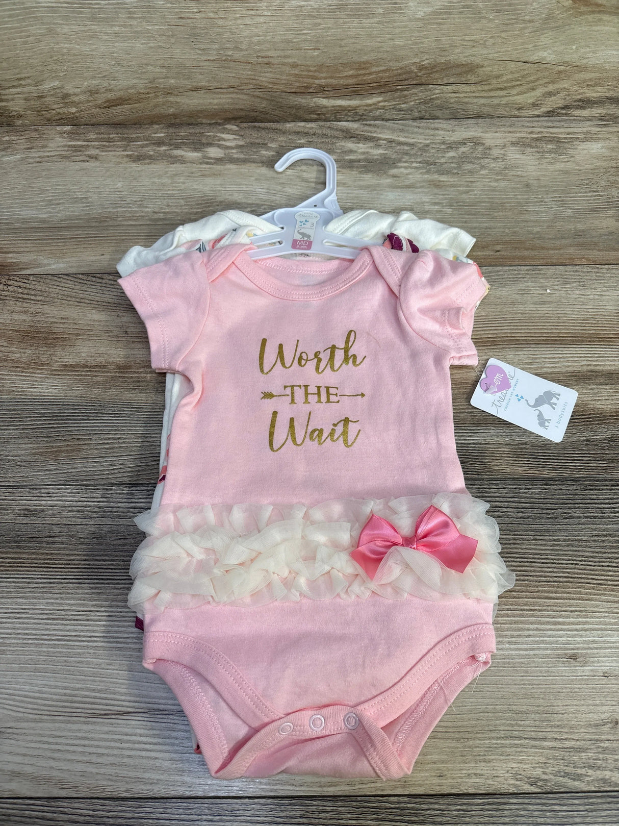 Flat lay of a 3-pack of baby bodysuits showing a pink bodysuit reading Worth the Wait with tulle ruffle and pink bow on wood background