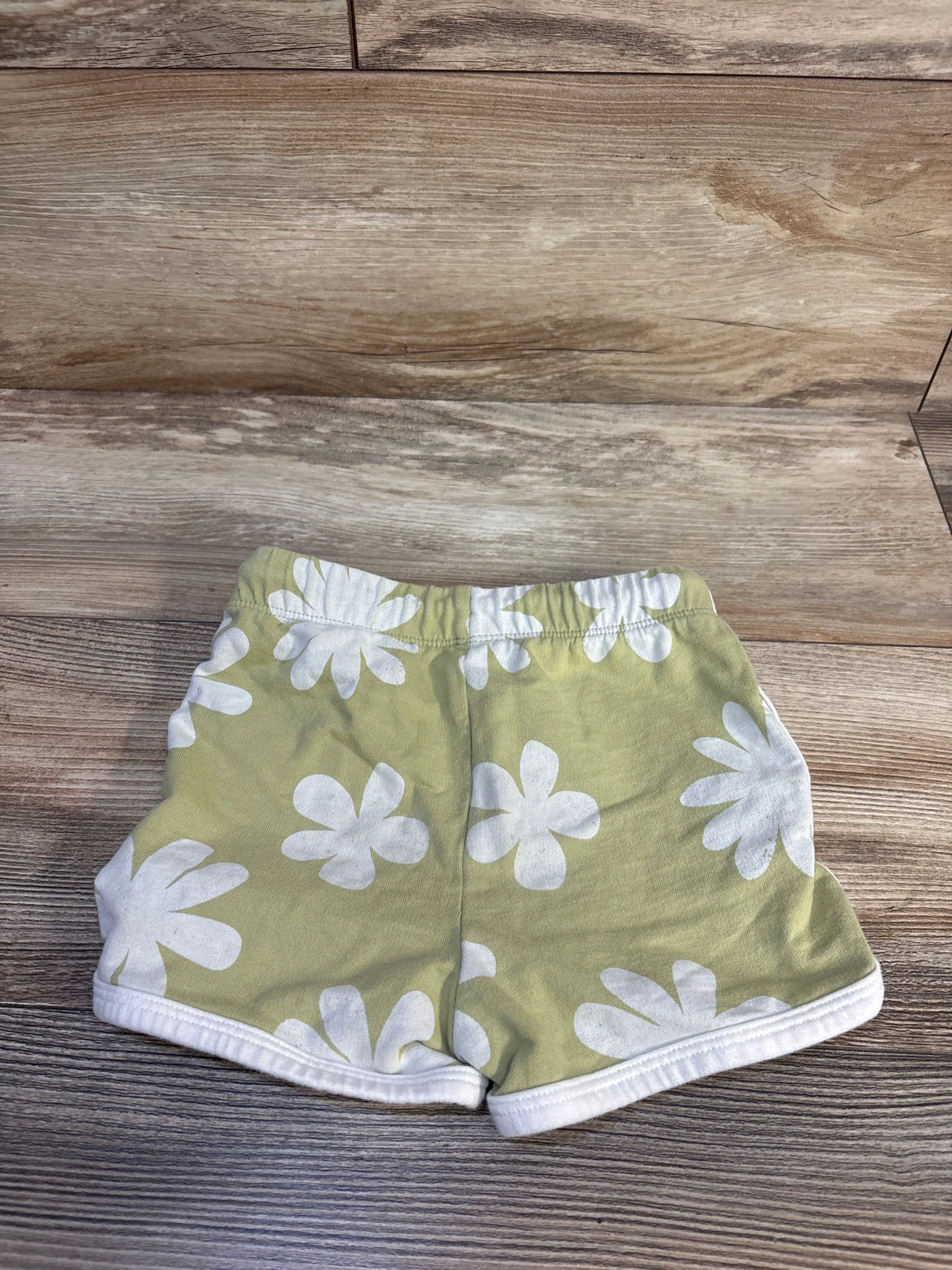 Green baby shorts with white daisy print, soft cotton fabric, laid on wood floor