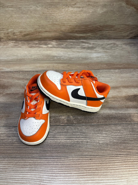 Angled view of Nike Dunk Low TD toddler sneakers showing black Swoosh, orange panels and laces on wood background