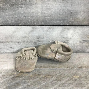 Front and side view of silver holographic soft-sole leather baby moccasins with fringe detailing on gray wooden background