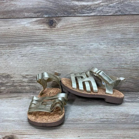 Three-quarter view of gold OshKosh Raquel toddler sandals with cork footbed, adjustable ankle strap, and cutout metallic upper on wood backdrop