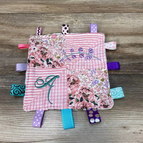Top-down of pink crinkle square with ribbon tabs showing embroidered initial A, purple vine and multicolored ribbon loops