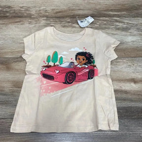 Front flat-lay of beige Children's Place toddler t-shirt showing a girl driving a pink car graphic with hearts and trees on a wood floor