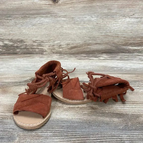 Three-quarter view of Zara baby fringe sandals in rust red, size 4C, showing braided ankle ties and fringe on wood-look background