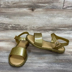 Angled view of Mel By Melissa toddler glitter jelly sandals showing gold glitter finish and ankle strap on wood floor