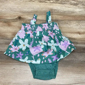 Flat lay of green floral baby bodysuit dress showcasing smocked bodice, shoulder straps, and three snap closures at crotch on wood background