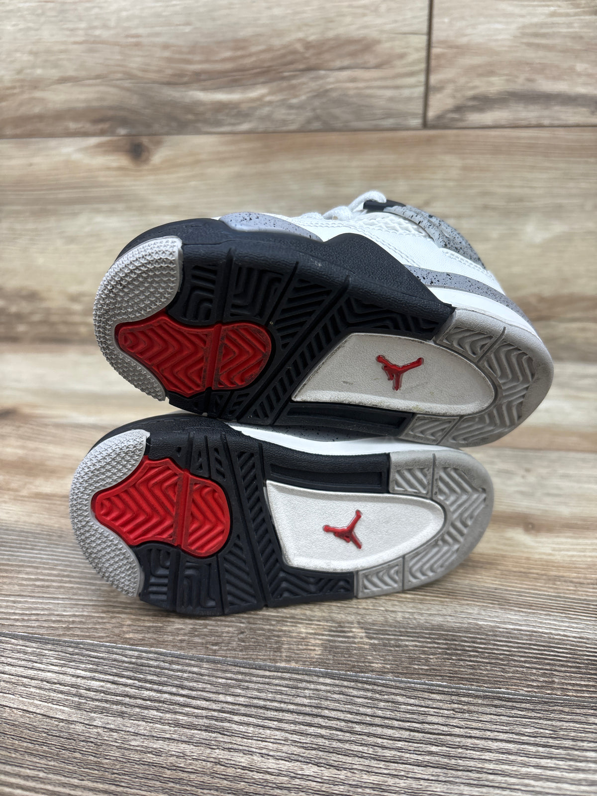 Outsole view of Air Jordan 4 Retro OG TD White Cement sneakers showcasing black, red, and gray tread with a wood floor background