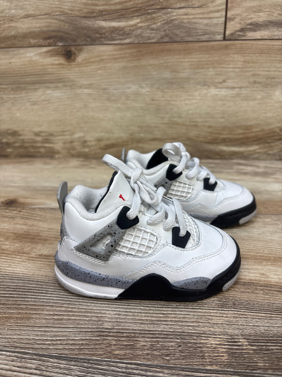 Three-quarter view of Air Jordan 4 Retro OG TD White Cement sneakers showing black accents and speckled gray midsoles on wood