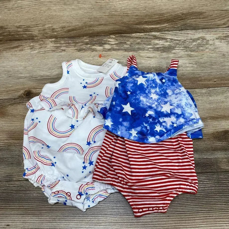 Flat lay of two 0–3M baby rompers showcasing a white rainbow-print romper with ruffle and a red-and-white striped romper with blue star top on wooden floor