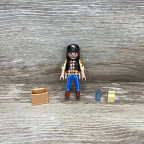 Front view of Playmobil 5665 dump truck driver figure with black helmet, plaid shirt, blue pants and three small accessories on wood background