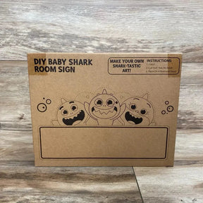 Front of Baby Shark DIY room sign box showing three cartoon sharks and a blank nameplate on a wood plank background